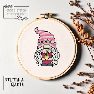 May include: Cross stitch pattern PDF of a pink and white gnome holding a yellow mug. The pattern is displayed in a wooden embroidery hoop.