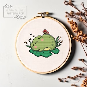 May include: Cross stitch pattern PDF of a green frog sleeping on a lily pad with a red mushroom on top of the pad. The frog has pink cheeks and is surrounded by green leaves.