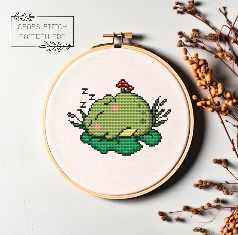 May include: Cross stitch pattern PDF of a green frog sleeping on a lily pad with a red mushroom on top of the pad. The frog has pink cheeks and is surrounded by green leaves.