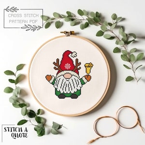 May include: Cross stitch pattern PDF of a Christmas gnome wearing a red hat with a snowflake, holding a bell and antlers. The pattern is displayed in a wooden embroidery hoop.