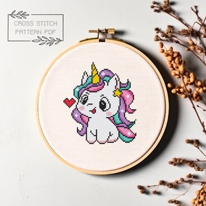 Puede incluir: Patrón de punto de cruz de un lindo unicornio de dibujos animados con un corazón rosa, una melena morada y azul y un cuerno amarillo. El unicornio está sentado sobre un fondo blanco.
