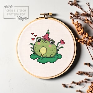May include: Cross stitch pattern PDF of a green frog wearing a pink flower on its head, sitting on a green lily pad with three red hearts floating above it.