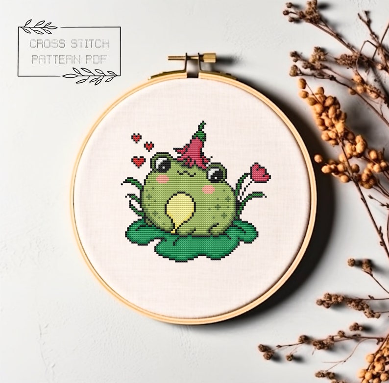 May include: Cross stitch pattern PDF of a green frog wearing a pink flower on its head, sitting on a green lily pad with three red hearts floating above it.