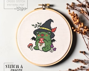 May include: Cross stitch pattern PDF of a green frog wearing a witch hat and holding a red mushroom. The frog has wings and is surrounded by mushrooms.