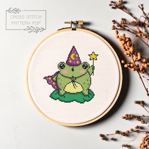 May include: A cross stitch pattern featuring a green frog wearing a purple wizard hat and holding a star wand. The frog is sitting on a green lily pad.