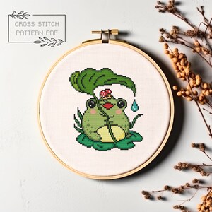 May include: Cross stitch pattern PDF of a green frog sitting under a large green leaf with a red mushroom on its head. The frog is holding the leaf with its hands and there is a blue raindrop falling from the leaf.