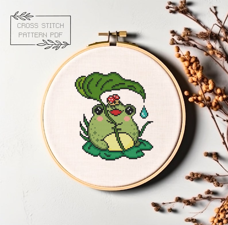 May include: Cross stitch pattern PDF of a green frog sitting under a large green leaf with a red mushroom on its head. The frog is holding the leaf with its hands and there is a blue raindrop falling from the leaf.