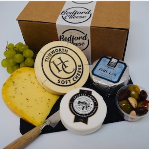 May include: A selection of cheeses from The Bedford Cheese Company, including Tunworth Soft Cheese, Perl Las, and Snowdonia Truffle Trove. The image shows a cheese knife, grapes, olives, and a black slate.