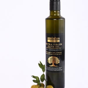 May include: A tall, dark green glass bottle of Andalus Premium Extra Virgin Olive Oil with a black cap and a black label featuring gold text and a gold tree illustration. Three green olives with small stems and leaves are placed at the base.