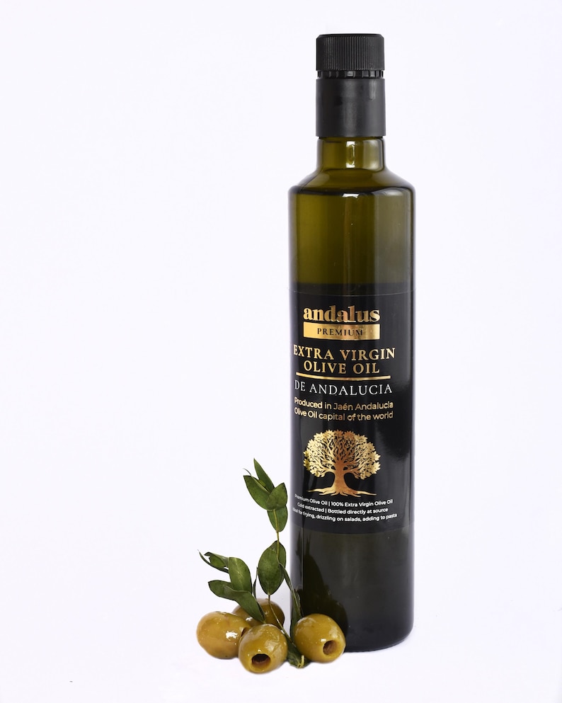 May include: A tall, dark green glass bottle of Andalus Premium Extra Virgin Olive Oil with a black cap and a black label featuring gold text and a gold tree illustration. Three green olives with small stems and leaves are placed at the base.