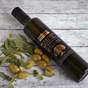 May include: A dark glass bottle of Andalus Premium Extra Virgin Olive Oil with a black cap, featuring a label with a gold tree illustration and text. Beside the bottle are several fresh green olives attached to small leafy branches.