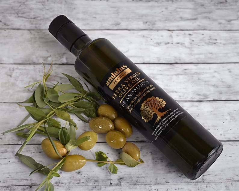 May include: A dark glass bottle of Andalus Premium Extra Virgin Olive Oil with a black cap, featuring a label with a gold tree illustration and text. Beside the bottle are several fresh green olives attached to small leafy branches.