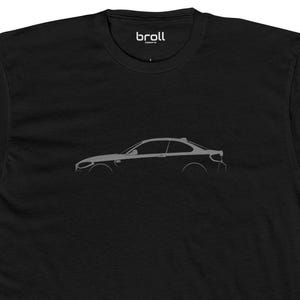 May include: Black t-shirt with a grey outline of a car on the front. The car is a BMW M2.