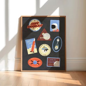 May include: A framed art print featuring a collection of colorful space-themed patches. The patches include text like "COPY MOON JOY" and "ARTEMIS", and images of rockets, astronauts, and planets. The frame is a light brown color.