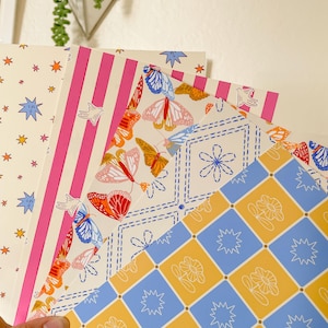 May include: A collection of patterned papers with various designs. One paper features pink stripes, another has a butterfly and floral pattern, and a third displays a star and floral pattern. The papers are held by a hand.