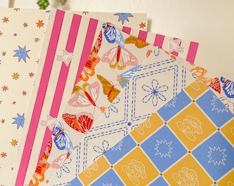 May include: A collection of patterned papers with various designs. One paper features pink stripes, another has a butterfly and floral pattern, and a third displays a star and floral pattern. The papers are held by a hand.