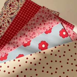 May include: A collection of patterned papers in various designs and colors. The papers feature floral, gingham, polka dot, and other decorative patterns in shades of red, pink, and cream. The papers are fanned out, showcasing the variety of designs.