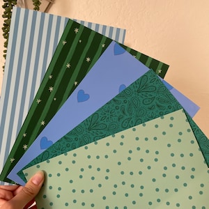 May include: A hand holds a fan of patterned paper sheets. The sheets display various designs, including stripes, stars, hearts, paisley, and polka dots, in shades of blue and green. The paper is likely for crafts or stationery.