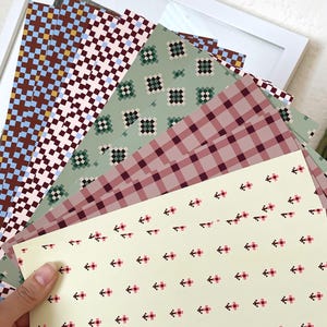 May include: A collection of rectangular paper sheets featuring various patterns including a light yellow sheet with small pink and black pixelated crosses, a mauve gingham check, a sage green pixelated floral design, and brown and blue grids.