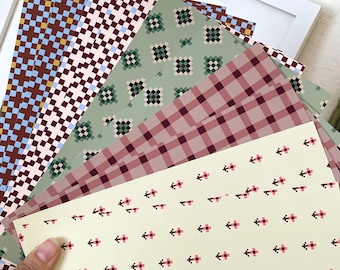 Op de afbeelding: Een verzameling rechthoekige vellen papier met diverse patronen, waaronder een lichtgeel vel met kleine roze en zwarte gepixelde kruisjes, een mauve ruitjespatroon, een saliegroen gepixeld bloemendesign en bruine en blauwe rasters.