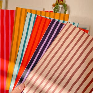 Pu&ograve; includere: Una mano tiene in mano un ventaglio di carte a righe colorate. Le carte presentano strisce verticali in vari colori, tra cui rosso, arancione, turchese, viola e rosa. Le carte sono a ventaglio, mostrando le diverse combinazioni di colori e motivi.