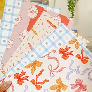 May include: A hand holds a stack of patterned papers. Designs include blue gingham, pink with cake illustrations, red with candles, and cream with ribbons in orange, pink, and purple. The papers are suitable for crafts, scrapbooking, or gift wrapping.