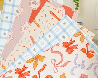 May include: A hand holds a stack of patterned papers. Designs include blue gingham, pink with cake illustrations, red with candles, and cream with ribbons in orange, pink, and purple. The papers are suitable for crafts, scrapbooking, or gift wrapping.