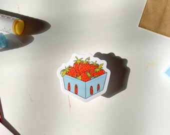 May include: A sticker featuring a cartoon illustration of ripe strawberries in a light blue basket. The sticker is on a white surface, with receipts and other stationery items visible in the background.