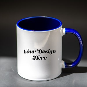 K&ouml;nnte beinhalten: Wei&szlig;er Keramikbecher mit blauem Innenraum und Henkel. Der Becher tr&auml;gt den Text "Your Design Here" in einer dekorativen Schriftart. Der Becher steht auf einer grauen Oberfl&auml;che mit dunklem Hintergrund.
