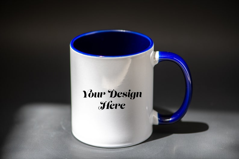 K&ouml;nnte beinhalten: Wei&szlig;er Keramikbecher mit blauem Innenraum und Henkel. Der Becher tr&auml;gt den Text "Your Design Here" in einer dekorativen Schriftart. Der Becher steht auf einer grauen Oberfl&auml;che mit dunklem Hintergrund.
