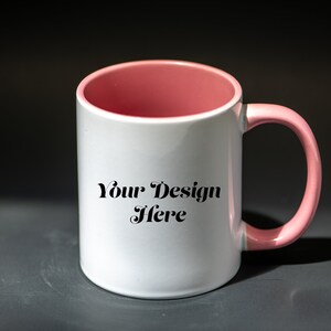 K&ouml;nnte beinhalten: Wei&szlig;er Keramikbecher mit rosa Innenraum und Henkel. Der Becher tr&auml;gt den Text "Your Design Here" in schwarzer Schrift. Der Becher steht vor einem dunkelgrauen Hintergrund.