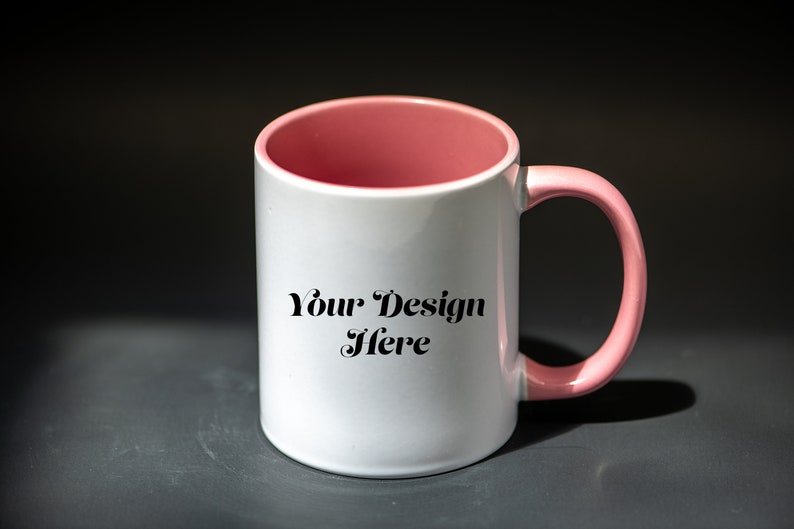 K&ouml;nnte beinhalten: Wei&szlig;er Keramikbecher mit rosa Innenraum und Henkel. Der Becher tr&auml;gt den Text "Your Design Here" in schwarzer Schrift. Der Becher steht vor einem dunkelgrauen Hintergrund.