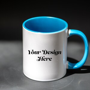 K&ouml;nnte beinhalten: Wei&szlig;er Keramikbecher mit hellblauem Innenraum und Henkel. Auf dem Becher steht der Text "Your Design Here". Der Becher steht auf einer grauen Oberfl&auml;che.