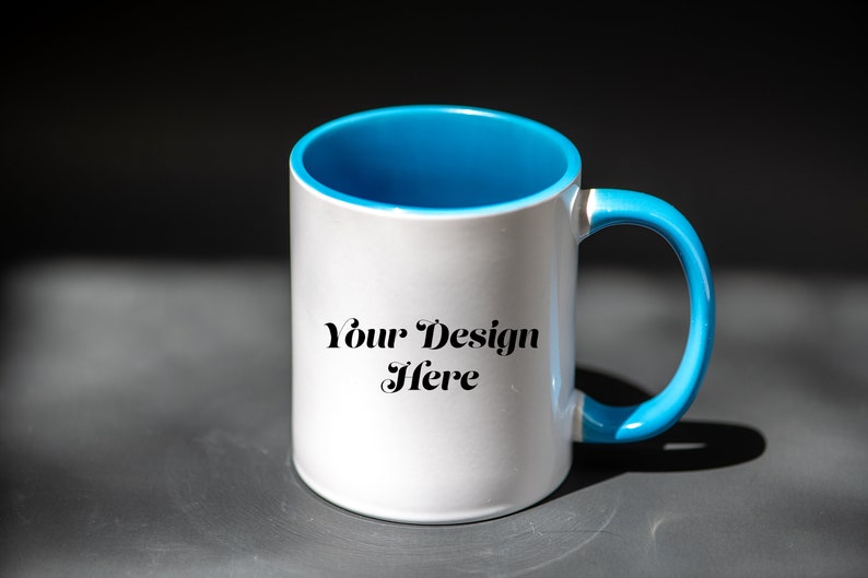 K&ouml;nnte beinhalten: Wei&szlig;er Keramikbecher mit hellblauem Innenraum und Henkel. Auf dem Becher steht der Text "Your Design Here". Der Becher steht auf einer grauen Oberfl&auml;che.