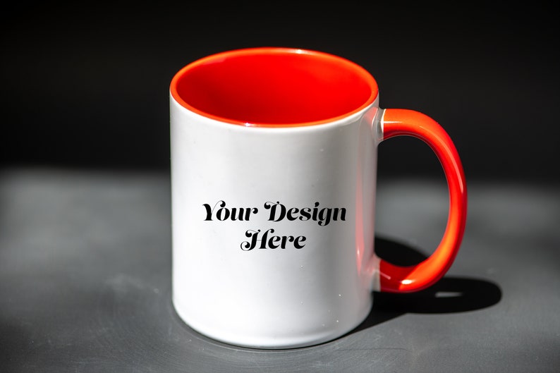 K&ouml;nnte beinhalten: Wei&szlig;er Keramikbecher mit leuchtend rotem Innenraum und Henkel. Der Becher tr&auml;gt den Text "Your Design Here" in schwarzer Schrift. Der Becher steht vor einem dunklen Hintergrund, der seine leuchtenden Farben und sein Design hervorhebt.