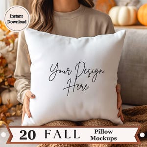 Op de afbeelding: Een wit vierkant kussen met de tekst "Your Design Here" in zwart script. Het kussen wordt voor een persoon gehouden. De afbeelding bevat ook de tekst "Instant Download" en "20 Fall Pillow Mockups". Pompoenen en herfstbladeren zijn op de achtergrond.