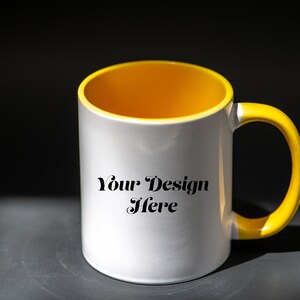 K&ouml;nnte beinhalten: Wei&szlig;e Keramiktasse mit gelbem Innenraum und Henkel. Auf der Tasse steht der Text "Your Design Here". Die Tasse steht auf einer dunkelgrauen Oberfl&auml;che mit schwarzem Hintergrund.