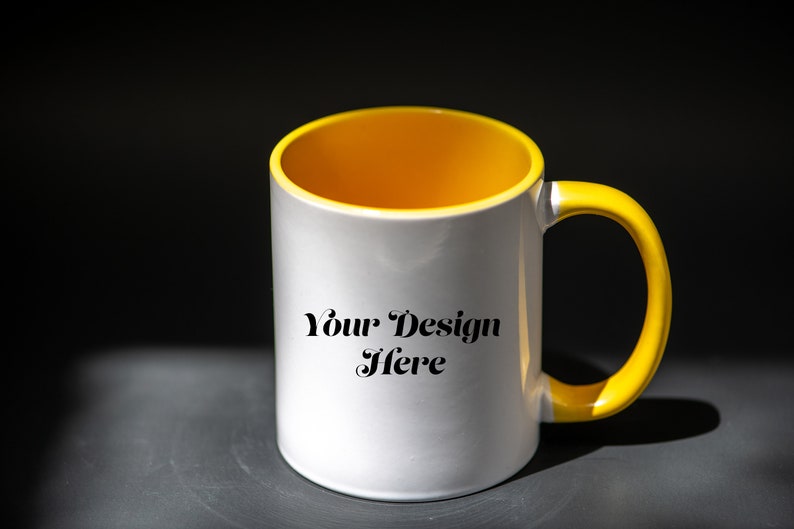 K&ouml;nnte beinhalten: Wei&szlig;e Keramiktasse mit gelbem Innenraum und Henkel. Auf der Tasse steht der Text "Your Design Here". Die Tasse steht auf einer dunkelgrauen Oberfl&auml;che mit schwarzem Hintergrund.