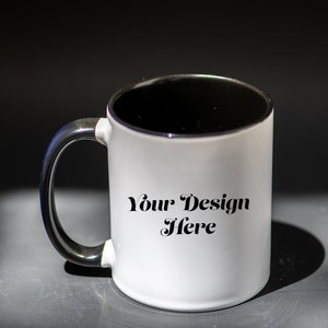 K&ouml;nnte beinhalten: Wei&szlig;er Keramikbecher mit schwarzem Innenraum und Henkel. Der Becher tr&auml;gt den Text "Your Design Here". Der Becher steht auf einer dunklen Oberfl&auml;che.