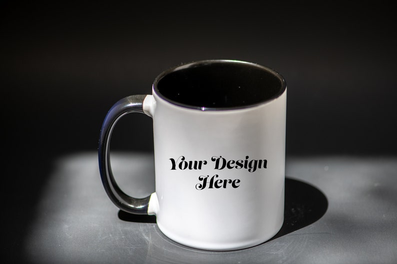 K&ouml;nnte beinhalten: Wei&szlig;er Keramikbecher mit schwarzem Innenraum und Henkel. Der Becher tr&auml;gt den Text "Your Design Here". Der Becher steht auf einer dunklen Oberfl&auml;che.