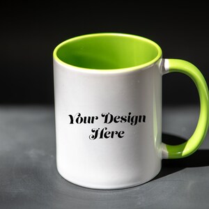 K&ouml;nnte beinhalten: Wei&szlig;er Keramikbecher mit gr&uuml;nem Innenraum und Griff. Der Becher tr&auml;gt den Text "Your Design Here" in schwarzer Schrift. Der Becher ist zylindrisch und steht auf einer dunkelgrauen Oberfl&auml;che.