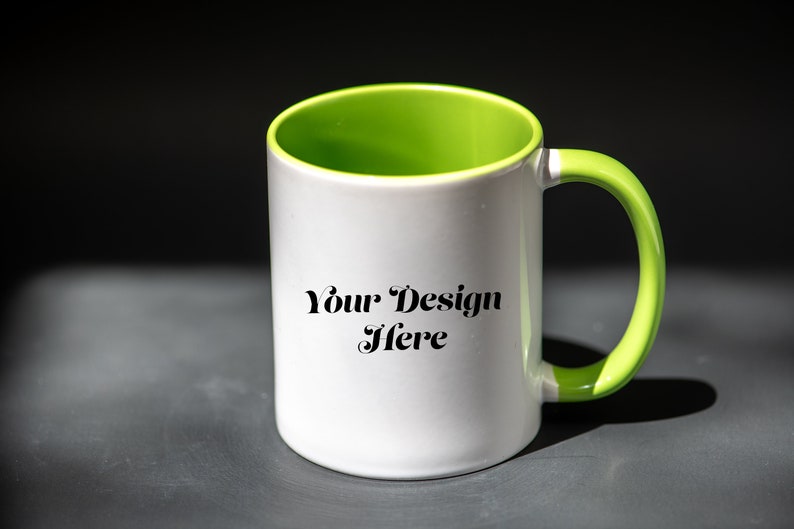 K&ouml;nnte beinhalten: Wei&szlig;er Keramikbecher mit gr&uuml;nem Innenraum und Griff. Der Becher tr&auml;gt den Text "Your Design Here" in schwarzer Schrift. Der Becher ist zylindrisch und steht auf einer dunkelgrauen Oberfl&auml;che.