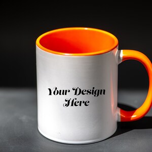 K&ouml;nnte beinhalten: Wei&szlig;er Keramikbecher mit orangefarbenem Innenraum und Henkel. Auf dem Becher steht der Text "Your Design Here". Der Becher steht auf einer dunkelgrauen Oberfl&auml;che.