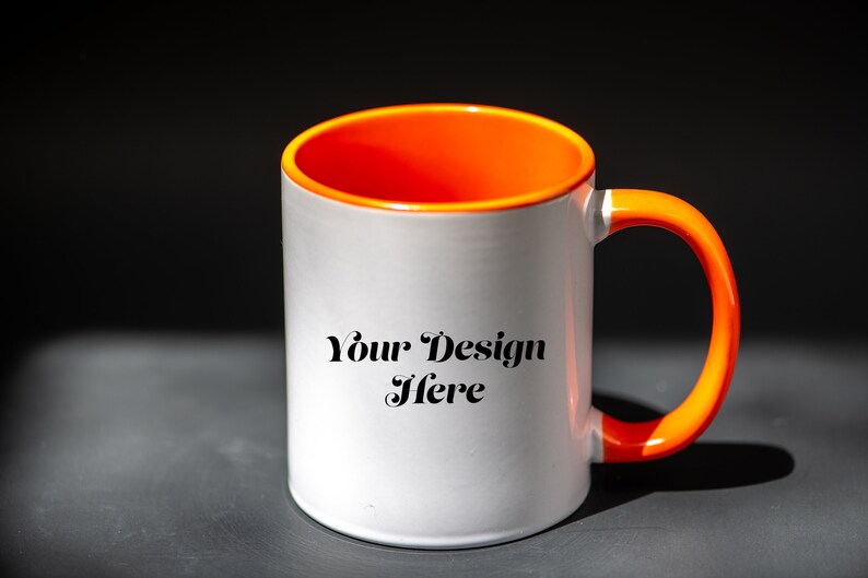 K&ouml;nnte beinhalten: Wei&szlig;er Keramikbecher mit orangefarbenem Innenraum und Henkel. Auf dem Becher steht der Text "Your Design Here". Der Becher steht auf einer dunkelgrauen Oberfl&auml;che.
