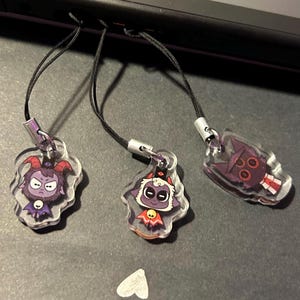 May include: Three clear acrylic charms with black cords. Each charm features a cartoon character with a dark purple and red color scheme. The charms are attached to a black device.