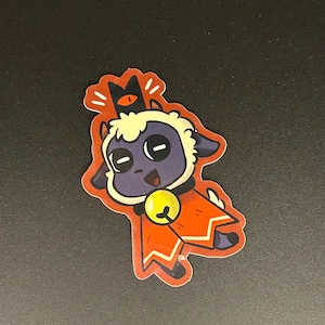 May include: A sticker featuring a cartoon lamb with a purple face, cream-colored wool, and a red cloak. The lamb has a yellow bell around its neck and a black crown. The sticker has a red border.