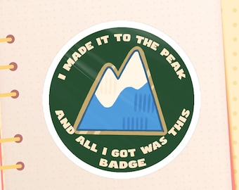May include: A round sticker with the text "I MADE IT TO THE PEAK AND ALL I GOT WAS THIS BADGE". The sticker features a mountain graphic with blue and white colors, set against a green background with a gold border.