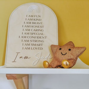 May include: A wooden sign with the text "I am Fun, I am Kind, I am Brave, I am Honest, I am Caring, I am Special, I am Confident, I am Strong, I am Smart, I am Loved" and the name "Judy" written below. A gold glitter star with a smiley face is sitting on the shelf next to the sign. A brown yarn sculpture of the word "hello" is on the shelf below the sign. A gray card with the text "You are magic" is on the shelf next to the yarn sculpture.