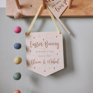 May include: A wooden sign with the text "Easter Bunny please stop here for Olivia & Mabel" hanging on a wooden rack with a yellow ribbon. The sign is decorated with bunny ears and a string of colourful felt balls hangs from the rack.