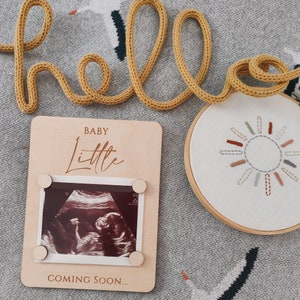 May include: A wooden photo frame with a white background and the text "BABY Little COMING SOON..." in brown lettering. The frame has a clear plastic insert to hold a sonogram photo.
